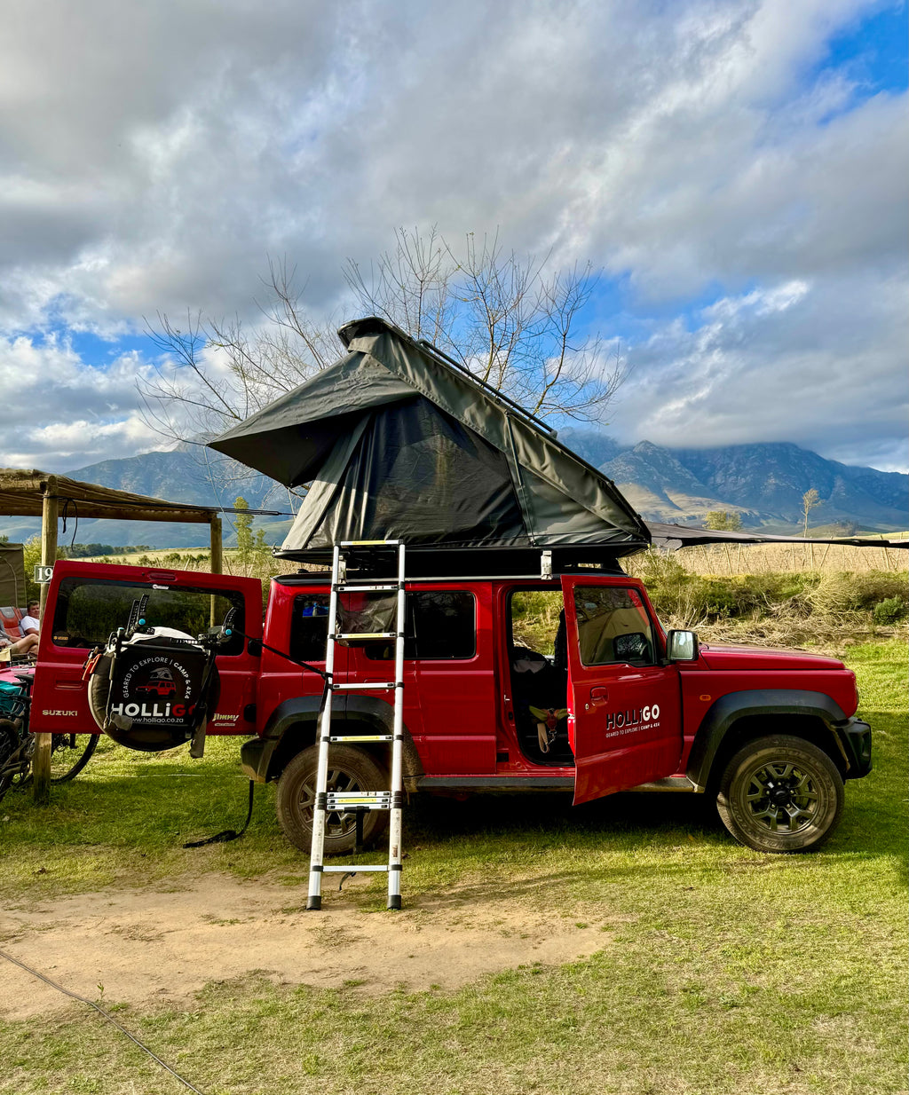 Red SUV with a rooftop tent in a natural setting with mountains and clouds.