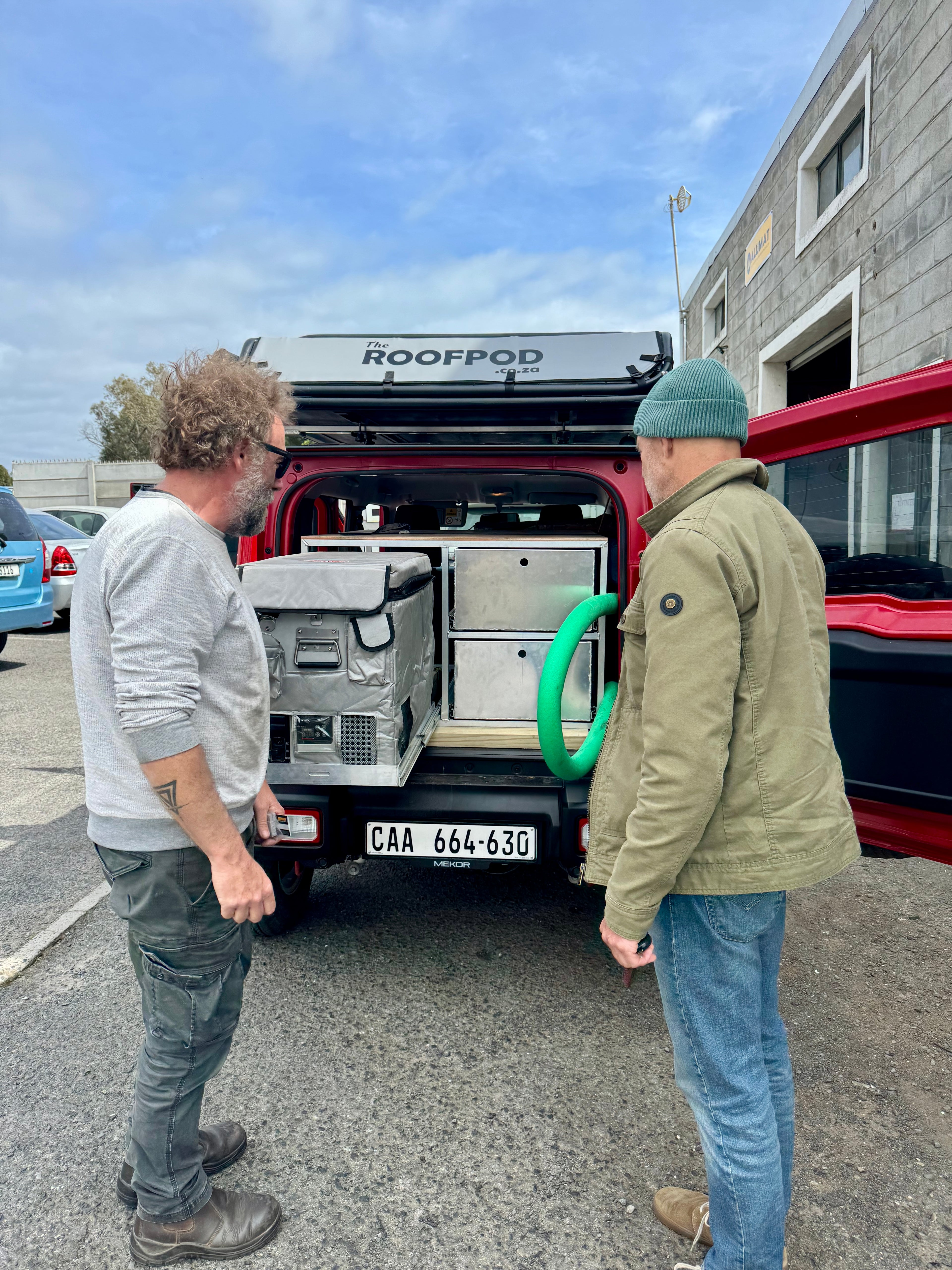 Two men standing next to a Suzuki Jimny with 'Roofpod' on the roof, in an outdoor setting.