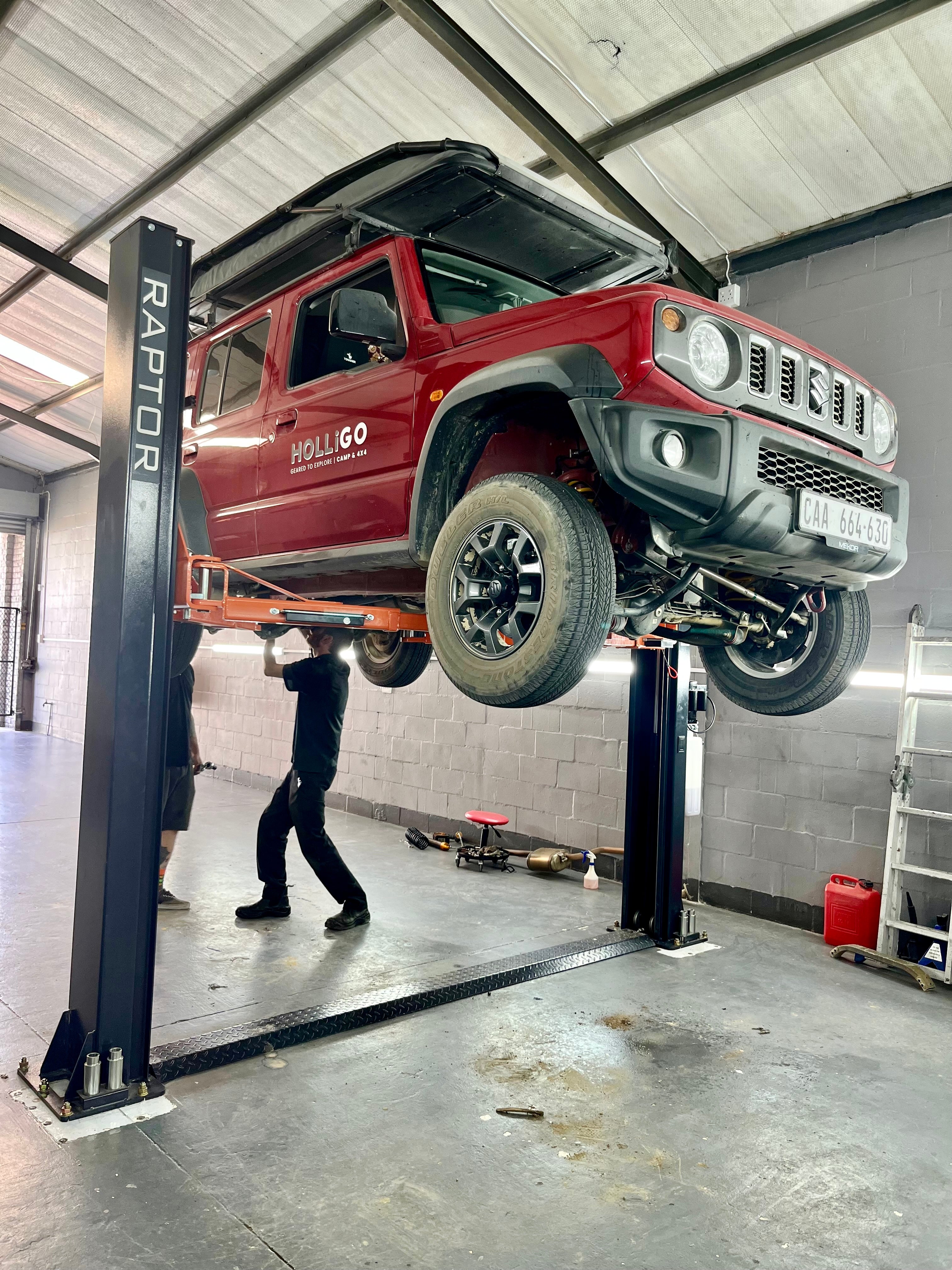 Red Suzuki Jimny on a lift in a garage with a person working underneath.