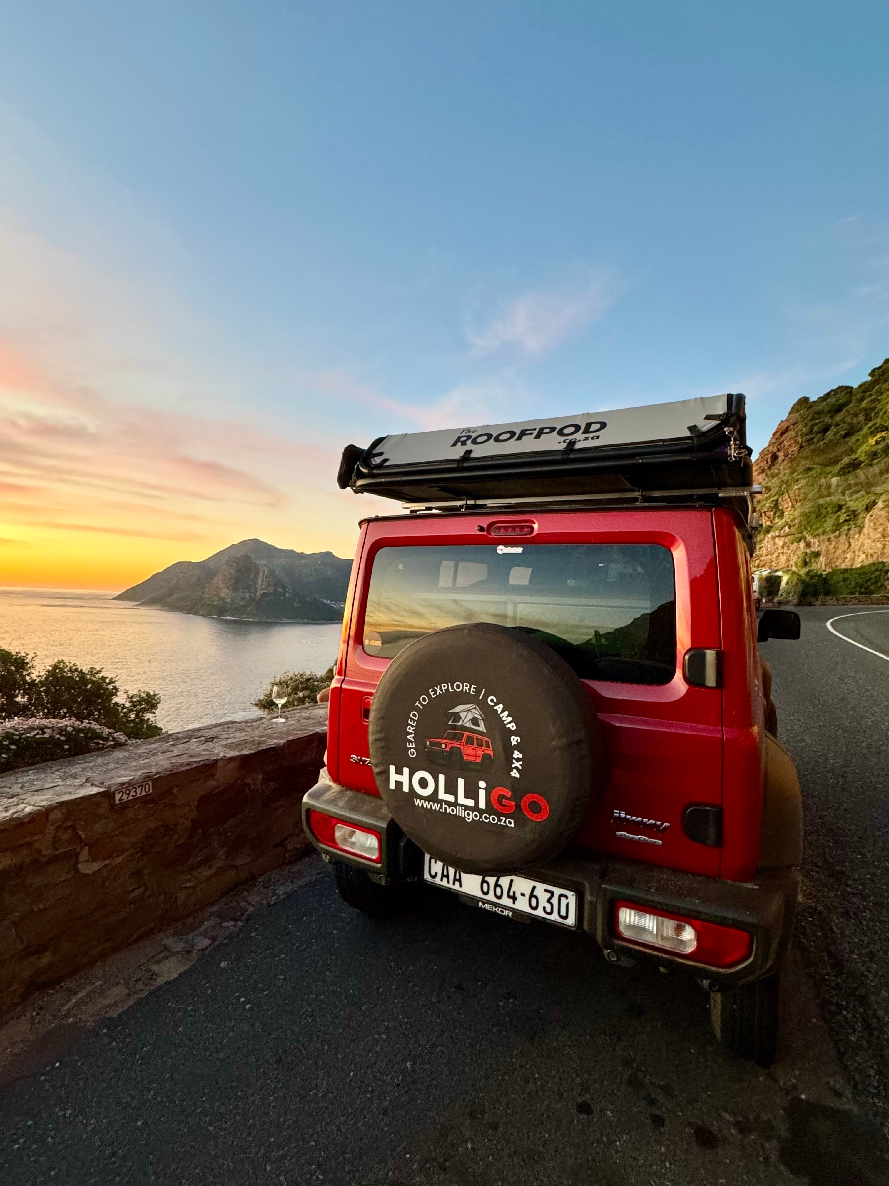 Red SUV with a spare tyre cover on a scenic road with mountains and sunset.